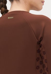 Ruda ilgomis rankovėmis palaidinė, dekoruota gėlių raštais ant rankovių ir šviesiai žalia Roxy logotipu prie kaklo. Švelni tekstūra.