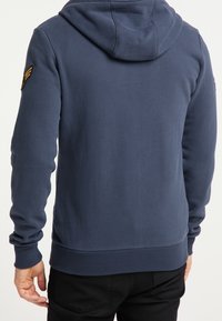 Granatowy hoodie wykonany z miękkiego materiału, z przednią kieszenią kangurką, ściągaczami przy mankietach oraz naszywką z logo na lewym ramieniu.