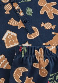 Tissu marine avec des bonhommes en pain d'épice, des bonbons et des motifs de fête en beige et vert. Comprend un col boutonné et un détail de taille élastique.