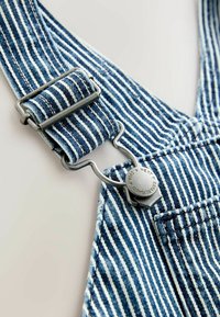 Blaue und weiße gestreifte Denim-Latzhose mit Metallbeschlägen, ausgestattet mit einer Clip-Schnalle und einem Knopfdetail auf dem Stoff.