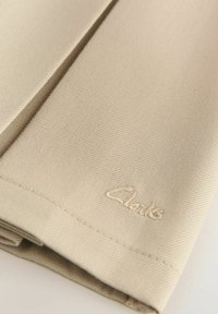 Primo piano di un tessuto beige piegato con tessitura fine e un sottile logo ricamato "Clarks" vicino al bordo.