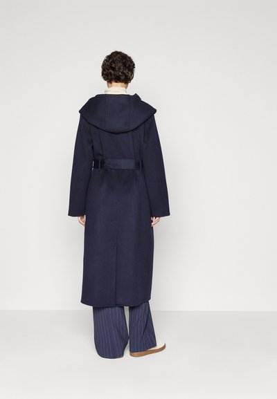 Lungo cappotto blu con un ampio cappuccio, vita regolata da una cintura e maniche larghe, abbinato a pantaloni a righe sottili. La texture appare liscia e ben rifinita.