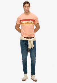 Lys orange bomulds t-shirt med røde og gule vandrette striber, der viser "Superdry" tekst. Sammen med blå jeans og en hvid sweater, der er bundet om taljen.