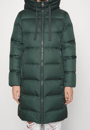 Długa ciemnozielona pikowana zimowa kurtka z przednim zamkiem błyskawicznym, kapturem, sznurkami i zapinanymi na zamek bocznymi kieszeniami, noszona na podartych białych dżinsach.