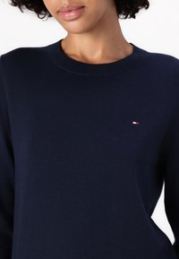 Granatowy sweter z okrągłym dekoltem wykonany z miękkiej tkaniny, z prążkowanymi mankietami. Na piersi małe logo w kolorach czerwonym, białym i niebieskim.