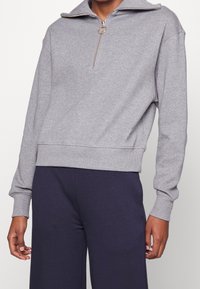 Pullover in pile grigio con collo a zip a un quarto e tirazip circolare. Presenta polsini e orlo a costine, con un discreto dettaglio del logo sul lato.