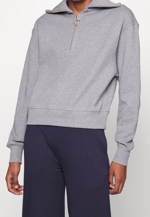 Grå fleece-pullover med en kvartdragkedja och en cirkulär dragkedjehanterare. Har ribbade ärmslut och nederkant, med en subtil logodetalj på sidan.