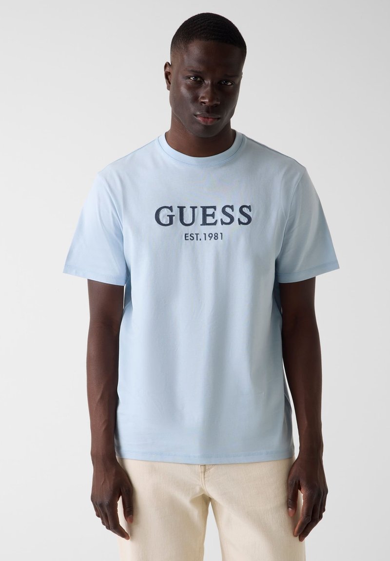 Homme noir portant un t-shirt bleu clair avec l'inscription "GUESS EST. 1981" en lettres foncées, associé à un pantalon beige sur un fond uni.