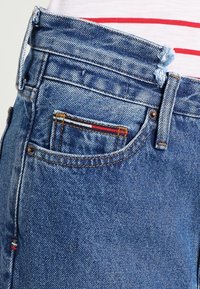 Blå denimjeans med en blekt textur, med en liten ficka med röda, vita och blå sömnadsdetaljer samt en metallknapp som stängning.