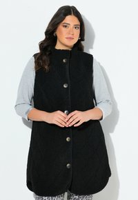 Nainen, jolla on yllään hihaton musta nappivaatteen fleeceliivi vaaleanharmaan pitkähihaisen paidan päällä ja leopardikuosiset housut, seisoo valkoista taustaa vasten.