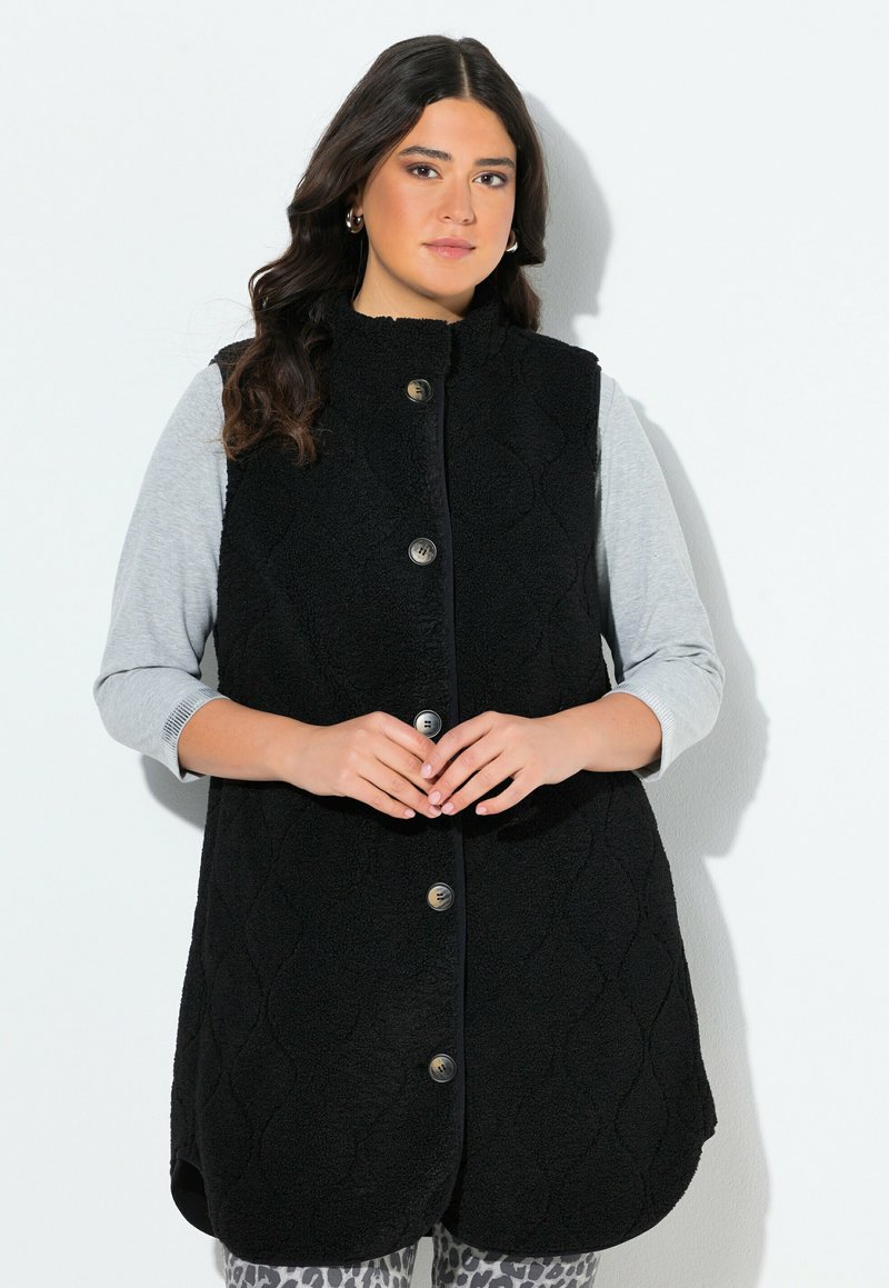 Kobieta w bezrękawnik czarnym zapinanym na guziki, nosząca jasnoszary długi rękaw, w legginsach w panterkę, stojąca na tle białym.