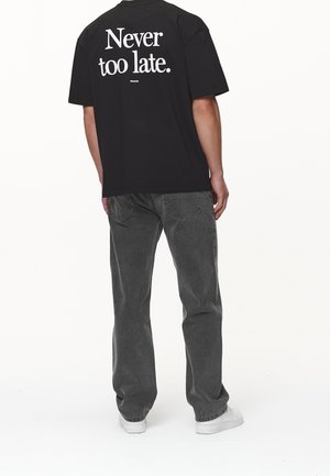 Camiseta de algodón negra con texto gráfico blanco "Nunca es demasiado tarde." en la parte posterior, combinada con jeans grises y zapatillas blancas.