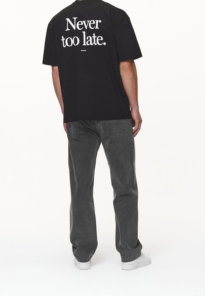 Camiseta de algodón negra con texto gráfico blanco "Nunca es demasiado tarde." en la parte posterior, combinada con jeans grises y zapatillas blancas.
