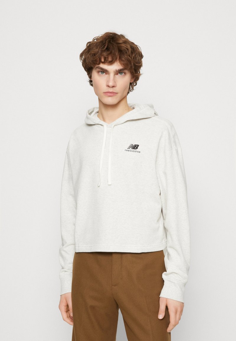 Haut court en molleton gris clair, fabriqué dans un tissu doux, comportant une poche avant, une capuche avec cordon de serrage, et un logo noir "New Balance" sur la poitrine.