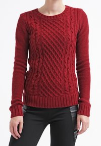 Röd stickad tröja med ett texturerat kabelmönster, rund halsringning och ribbade ärmslut och nederkant. Materialet verkar tjockt och varmt.
