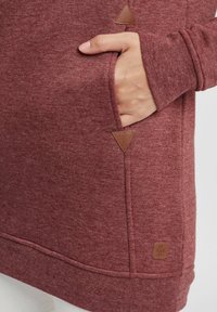 Haut à capuche bordeaux en tissu texturé, avec une poche latérale, des accents en cuir triangulaires et un ourlet cousu.
