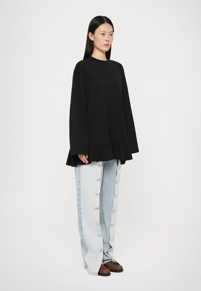 Fekete, oversized pulóver fodros aljjal, világoskék, széles szárú farmernadrággal, oldalsó gombokkal párosítva. A modellen barna slip-on cipők vannak.