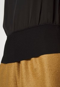 Top negro con cinturilla fruncida y dobladillo acanalado, combinado con una tela dorada de textura suave en la parte inferior, creando un efecto de capas contrastantes.