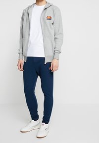 Felpa grigia con zip e logo, t-shirt bianca sotto, pantaloni jogger blu navy e sneakers bianche. Design semplice, vestibilità casual e materiale morbido.