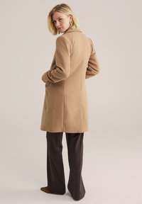 Cappotto di lana color cammello con vestibilità sartoriale, maniche lunghe e spacco sul retro. Abbinato a pantaloni a gamba larga marrone scuro.