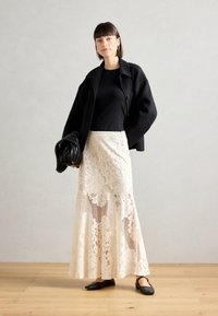Black wool blazer, fitted black top, cream lace maxi skirt with floral and mesh patterns, black flat shoes, holding a black textured bag.