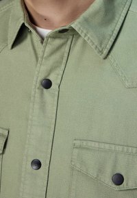 Camicia button-up verde con texture e bottoni neri, caratterizzata da due tasche frontali e un colletto classico, mostrata dalla vita in su.