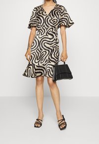 Krémové šaty ve stylu wrap s černým abstraktním vzorem, krátkým rukávem a volánkovým spodním lemem, doplněné černou kabelkou a černými sandály na podpatech.