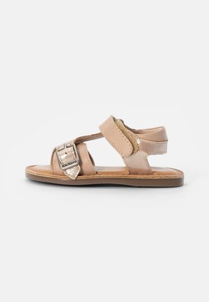 Sandalia de cuero marrón con correas ajustables, detalle de hebilla metálica, suela plana y diseño minimalista. Presenta un brillo sutil y una textura suave.