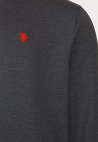 Camiseta de manga larga gris hecha de algodón suave con una textura sutil. Presenta un pequeño logotipo rojo bordado en el área del pecho izquierdo.