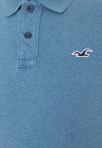 Polo triko z texturované tmavě modré látky má klasický límeček, tři černé knoflíky a malé bílé a černé logo na hrudi.