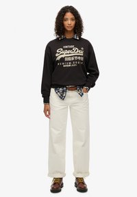 Zwart sweatshirt met "SuperDry"-logo, crème wijde broek, gelaagd geruit overhemd dat om de taille is geknoopt, en bruine veterlaarzen.