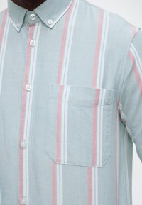 Ανοιχτό μπλε πουκάμισο με κάθετες λευκές και ροζ ρίγες, κουμπωτό μπροστινό μέρος, γιακά με κουμπιά και τσέπη στο στήθος στην αριστερή πλευρά.