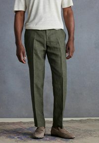 Olive green linen trousers with a straight cut, featuring a textured fabric and pressing crease, paired with light gray suede loafers.