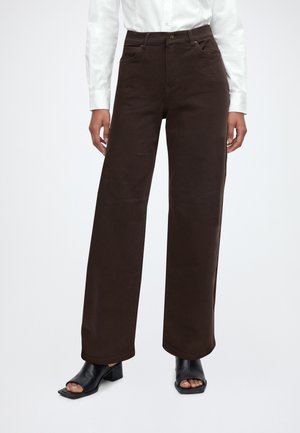 Personne portant un pantalon large marron foncé, une chemise blanche à manches longues et des chaussures à talons ouvertes noires, debout sur un fond blanc.