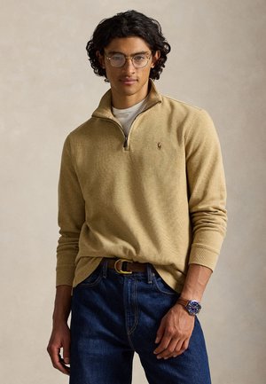 Young man with curly dark hair wearing round glasses, beige quarter-zip sweater, blue jeans, and a brown leather watch with a blue face.