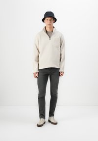 Krém színű bolyhos pulóver félcipzárral és elülső zsebbel; sötétszürke, slim-fit farmernadrággal és bézs sportcipővel párosítva. Sötét kalapban viselve.