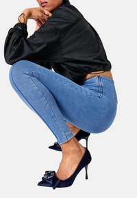 Marineblauwe hoge hakken met een ribbelstructuur, voorzien van een decoratieve strik en versieringen, gecombineerd met lichtblauwe skinny jeans.