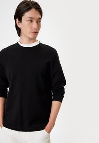 Czarna bawełniana bluza z długimi rękawami, z okrągłym dekoltem i prążkowanymi mankietami, nałożona na białą koszulkę. Minimalistyczny i gładki materiał.