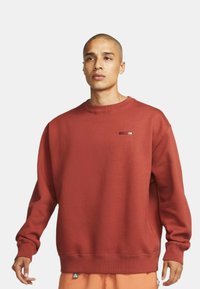 Czerwona bluza z okrągłym dekoltem i ściągaczami na rękawach oraz u dołu, z małą naszywką z logo na piersi. Miękki materiał o luźnym kroju.