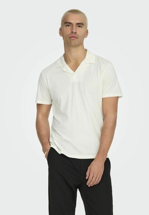Polo à manches courtes, de couleur crème, en tissu doux, avec un col en V et de subtiles accents au niveau du col. Associé à un pantalon noir.
