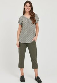 Grøn mønstret kortærmet bluse med V-hals, parret med olivenfarvede cropped bukser med knapdetaljer ved kanten; sorte slip-on sko.
