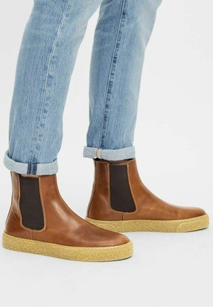 Bottes Chelsea en cuir marron avec panneaux latéraux élastiques noirs, portées avec un jean bleu clair retroussé sur fond blanc.
