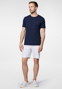 Granatowa koszulka z okrągłym dekoltem i krótkim rękawem, z małym czerwonym logo na klatce piersiowej. Połączona z białymi szortami i białymi sneakersami.