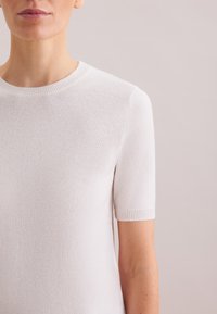 Maglione a maniche corte di colore chiaro, realizzato in tessuto morbido, con scollo arrotondato e polsini a coste per una texture aggiuntiva.