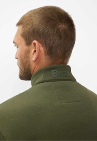 Pullover verde scuro con colletto alto, realizzato in un tessuto testurizzato e con un logo "Brax" in rilievo sul colletto posteriore. Finitura liscia.