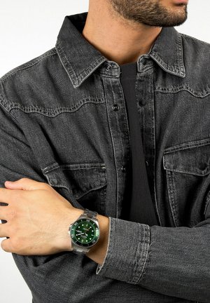Orologio in acciaio inossidabile con quadrante verde, marcatori argentati e funzione data, indossato su una mano. Giacca in denim grigio scuro con bottoni a pressione.