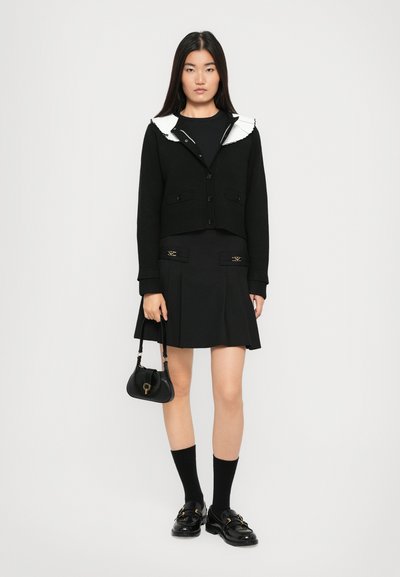 Cardigan nero tagliato con colletto bianco, gonna plissettata nera, calze al ginocchio e mocassini neri. Piccola borsa nera con dettagli dorati.