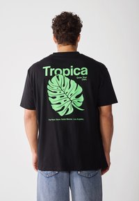 Svart T-skjorte med et grønt monstera-blad grafikk og teksten "Tropica" og "Grow Your Own" på baksiden, i bomullsmateriale.
