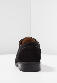 Scarpa da uomo in pelle scamosciata nera con punta rotonda e tacco basso, caratterizzata da un'elegante finitura in pelle liscia lungo il retro e una suola piatta e testurizzata.