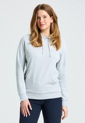Una mujer con una sudadera con capucha de color gris claro y pantalones azul marino posa sobre un fondo liso y claro.